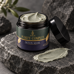A jar of AEMiLife Purity Complex Detox Mask with its black lid leaning against it, set on a dark gray stone slab with a smear of the light green mask in front. The background consists of dark rocks and soft-focus green leaves.