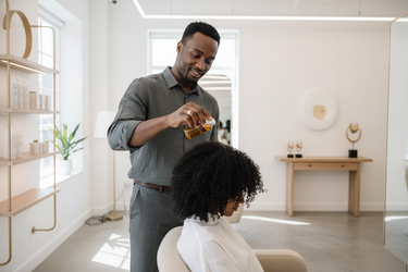 Black British-Ghanaian male stylist in his mid-30s, confident yet understated. He’s standing behind a salon chair with one hand in motion, applying oil to a client’s hair. The setting is bright, modern, and minimal — neutral tones, soft gold accents. His expression should feel warm, focused, and quietly proud — a craftsman in his element.